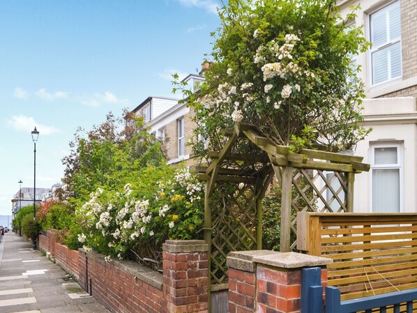 A residential street with brick walls, a wooden trellis with white flowers, and a bench.