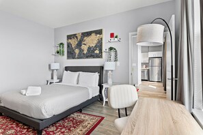 Een moderne slaapkamer met een groot bed, nachtkastjes, een stoel, een vloerkleed en een keuken zichtbaar door een geopende deur.
