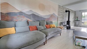 A modern living room with a grey sofa, colorful cushions, a large abstract mural, a stone wall, a black wood-burning stove, and a small wooden table. - The Villa in the country (PARIGNE)