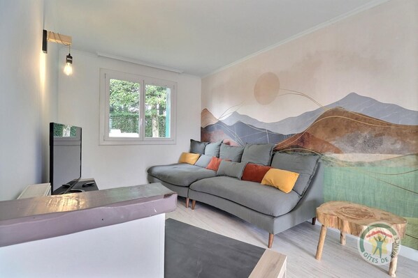 A modern living room with a grey sofa, a flat-screen TV, a window with a view of greenery, and a mural of mountains.