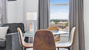 A modern living room with a grey sofa, a round glass table, and two chairs. A lamp is on the table, and the room has a large window with curtains. - Charming 1BR Near Encore xphu (Everett)