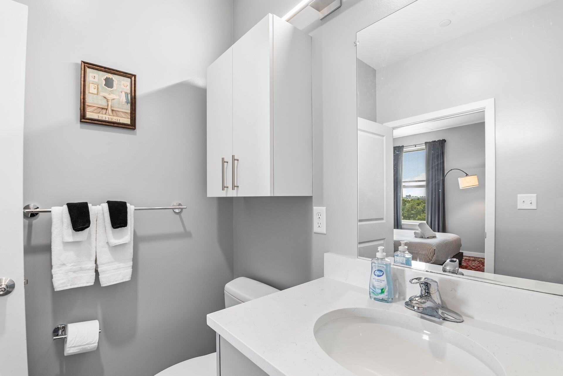 A modern bathroom with a white sink, a mirror, and a toilet. There is a towel rack with towels and a framed picture on the wall.