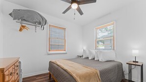 Un dormitorio con cama, cómoda de madera, ventilador de techo y dos ventanas con persianas.