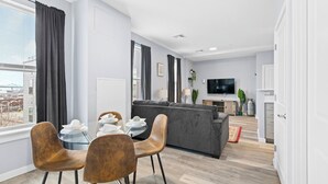 Een moderne woonkamer met een grijze bank, een eettafel met stoelen, een televisie en een uitzicht op de stad door grote ramen.