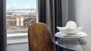 Een kamer met uitzicht op een brug en industriële gebouwen. Er staat een stoel en een tafel met een witte schaal en bord.