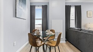 Een moderne eethoek met een glazen tafel en vier stoelen, een grijze bank en een stadszicht door de ramen.