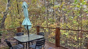 A wooden deck with a glass table and chairs, surrounded by a metal fence and trees with autumn leaves. - Riverfront | Trail & Kayak | Downtown & State Park (Mentone)