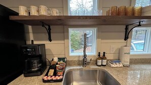 A kitchen with a stainless steel sink, a coffee maker, and a tray of food items. There are shelves above the sink with cups and a bottle of hand soap. - Riverfront | Trail & Kayak | Downtown & State Park (Mentone)