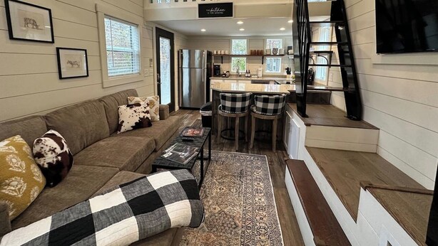 A cozy living room with a sofa, coffee table, and a rug. There is a kitchen area with a bar stool and a TV mounted on the wall. - Riverfront | Trail & Kayak | Downtown & State Park (Mentone)