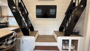 A modern kitchen with a central island, a built-in TV, and a staircase with black railings. - Riverfront | Trail & Kayak | Downtown & State Park (Mentone)