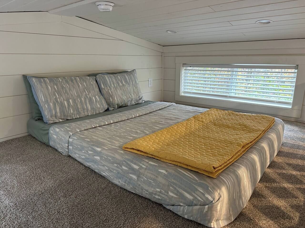 A bedroom with a bed, a window with blinds, and a carpeted floor.