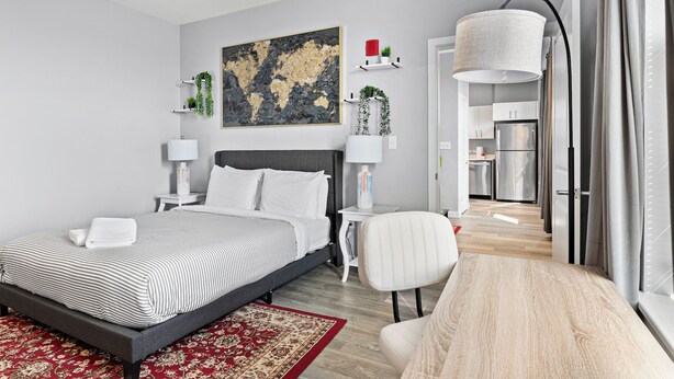 A modern bedroom with a large bed, bedside tables, a chair, a rug, and a kitchen visible through an open door. - Pleasant 1BR Near Encore Boston y69k (Everett)