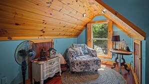 A cozy attic bedroom with a wooden ceiling, a bed with floral bedding, a dresser, a fan, and a window with a view of greenery.