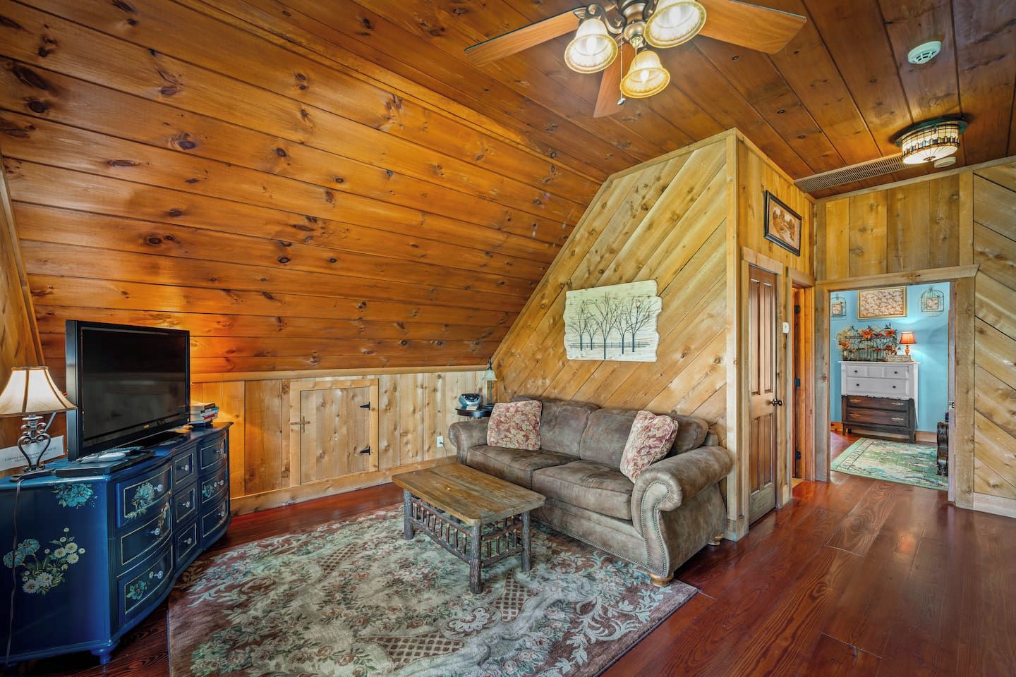 A cozy cabin interior with a wooden ceiling, a television on a dresser, a sofa with cushions, and a patterned rug.