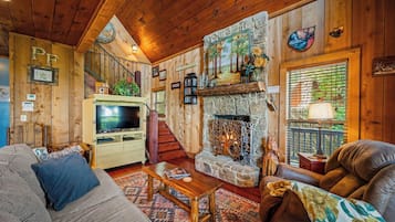 A cozy log cabin interior with a stone fireplace, wooden walls, a television, and a staircase.