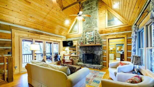 A cozy log cabin interior with a stone fireplace, wooden floors, and a large window overlooking a deck. - Serene Getaway | Hot Tub & Mountain Views (Mentone)