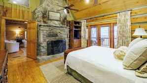 A cozy bedroom with a stone fireplace, wooden ceiling, and a large bed with decorative pillows. - Serene Getaway | Hot Tub & Mountain Views (Mentone)
