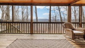 A covered porch with a wicker chair, wooden deck, and a view of trees and mountains. - Serene Getaway | Hot Tub & Mountain Views (Mentone)