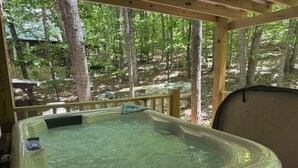 Un jacuzzi en un área boscosa con una terraza de madera y una zona de estar cubierta.
