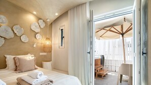 A bedroom with a bed, pillows, a bedside table, a wall with decorative elements, and a view of an outdoor seating area.