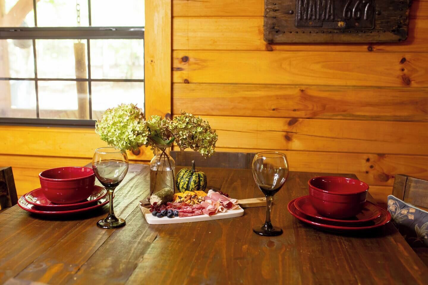 Una mesa de madera con platos rojos, copas de vino, un jarrón con flores y una bandeja de frutas.