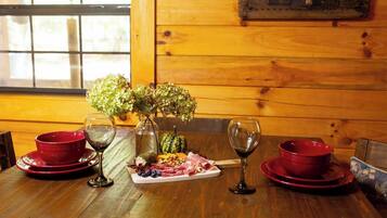 Una mesa de madera con platos rojos, copas de vino, un jarrón con flores y una bandeja de frutas.