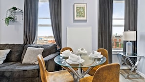 A modern living room with a gray sofa, a glass dining table with chairs, and a view of the city through large windows. - 1BR Near Encore Boston 45te (Everett)