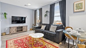A modern living room with a grey sofa, a TV, and a dining area with a glass table and chairs. - 1BR Near Encore Boston 45te (Everett)