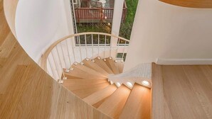 A spiral staircase with wooden steps and white railings, leading to a balcony with a view of greenery and a deck. - Crow's Nest Condo 3BR Downtown (Anchorage)