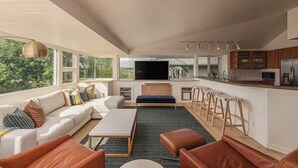 A modern living room with a sofa, ottoman, and a TV. There is a kitchen area with a bar and stools. - Crow's Nest Condo 3BR Downtown (Anchorage)