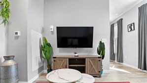 A modern living room with a flat-screen TV mounted on the wall, a wooden entertainment unit, two marble-topped coffee tables, a red patterned rug, and large green plants. - Nice 1BR Near Encore Boston tl51 (Everett)