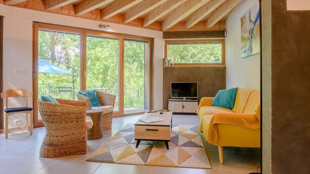 Una sala de estar moderna con techo de madera, ventanas amplias, un sofá amarillo, sillas de mimbre y un televisor.