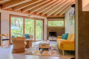 Un salon moderne avec un plafond en bois, de grandes fenêtres, un canapé jaune, des fauteuils en osier et un téléviseur.