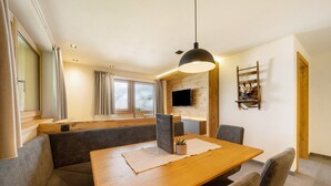 Un comedor moderno con una mesa de madera, sillas grises tapizadas, una lámpara colgante negra y un televisor montado en una pared de madera.