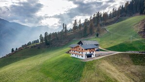 Una casa con techo inclinado, rodeada por un campo verde y árboles, con montañas al fondo.