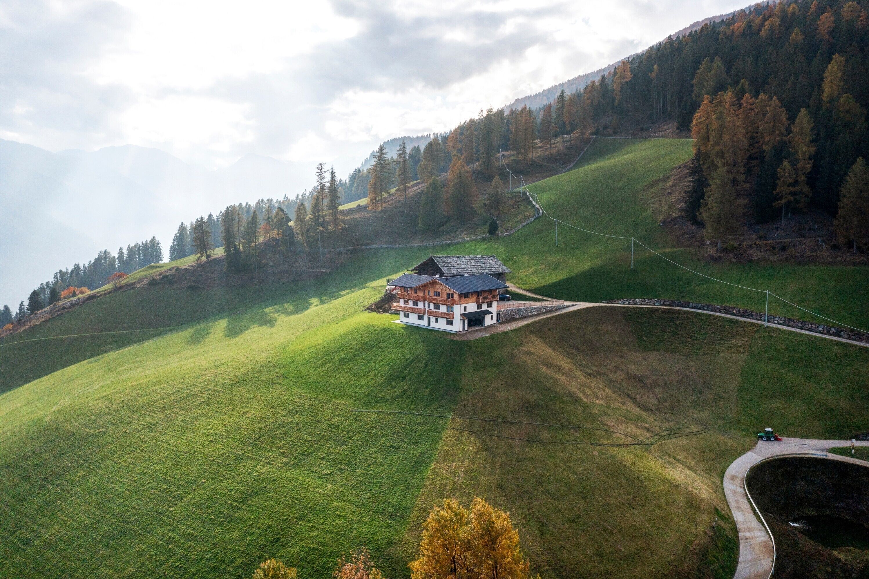 Ein Haus mit geneigtem Dach, umgeben von einem grünen Feld und Bäumen, mit einer kurvigen Straße in der Nähe.