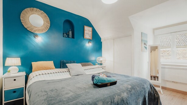 A bedroom with a blue accent wall, a round mirror, a bed with a blue blanket, a bedside table with a lamp, and a window with a view of a building. - Apartment Between Land and Sea – Peaceful Retreat Near Saint-Malo & Mont-Saint-Michel (Baguer-Pican)