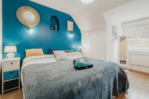 A bedroom with a blue accent wall, a round mirror, a bed with a blue blanket, a bedside table with a lamp, and a window with a view of a building.