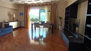 Een moderne woonkamer met een blauwe bank, een eethoek met een tafel en stoelen, een tv op een meubel en een groot raam met gordijnen.