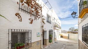 Una estrecha calle adoquinada con edificios blancos, una maceta roja con plantas, y un cartel que indica una zona rural.