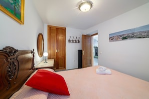 A bedroom with a bed, a red pillow, a wooden dresser, a mirror, a coat rack, and a painting on the wall.