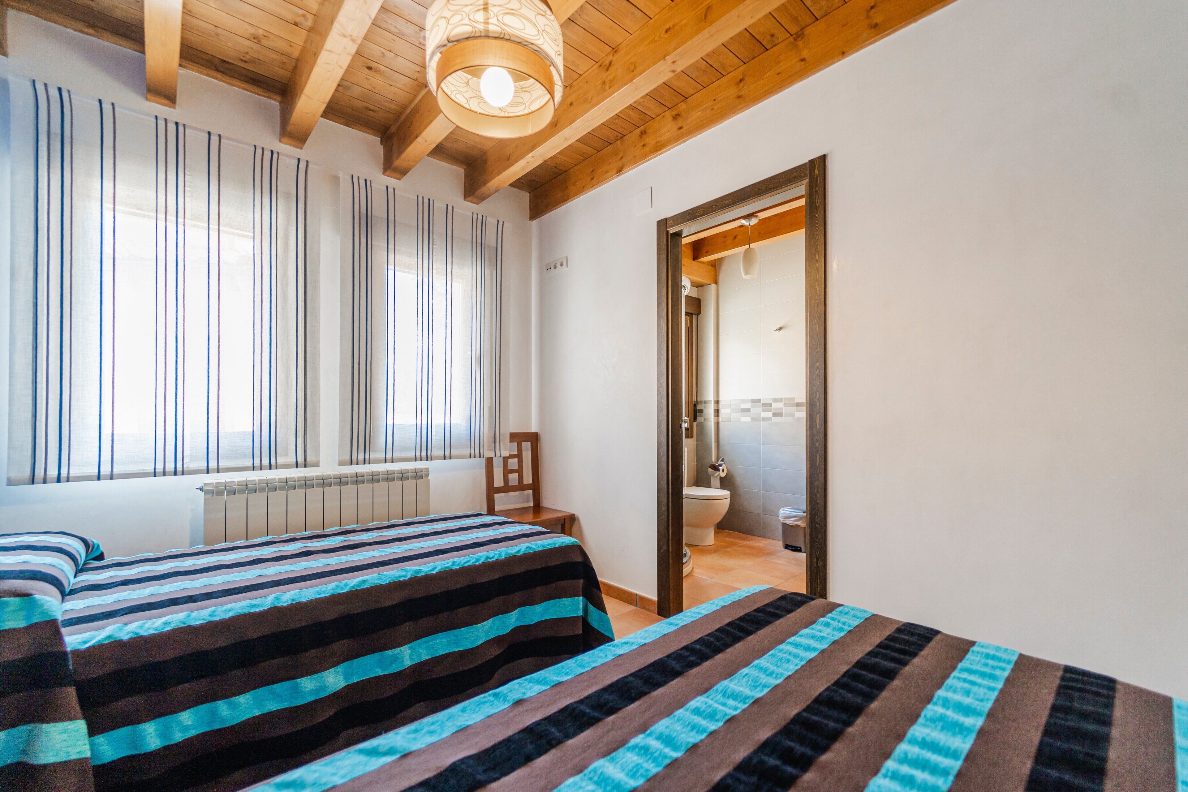 Een slaapkamer met twee bedden, een houten plafond, een raam met streepcurtains en een deur naar een badkamer.