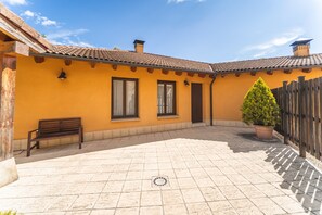 Une cour avec un sol carrelé, un banc en bois, une plante en pot et un bâtiment doté d’un toit en tuiles et de murs jaunes.