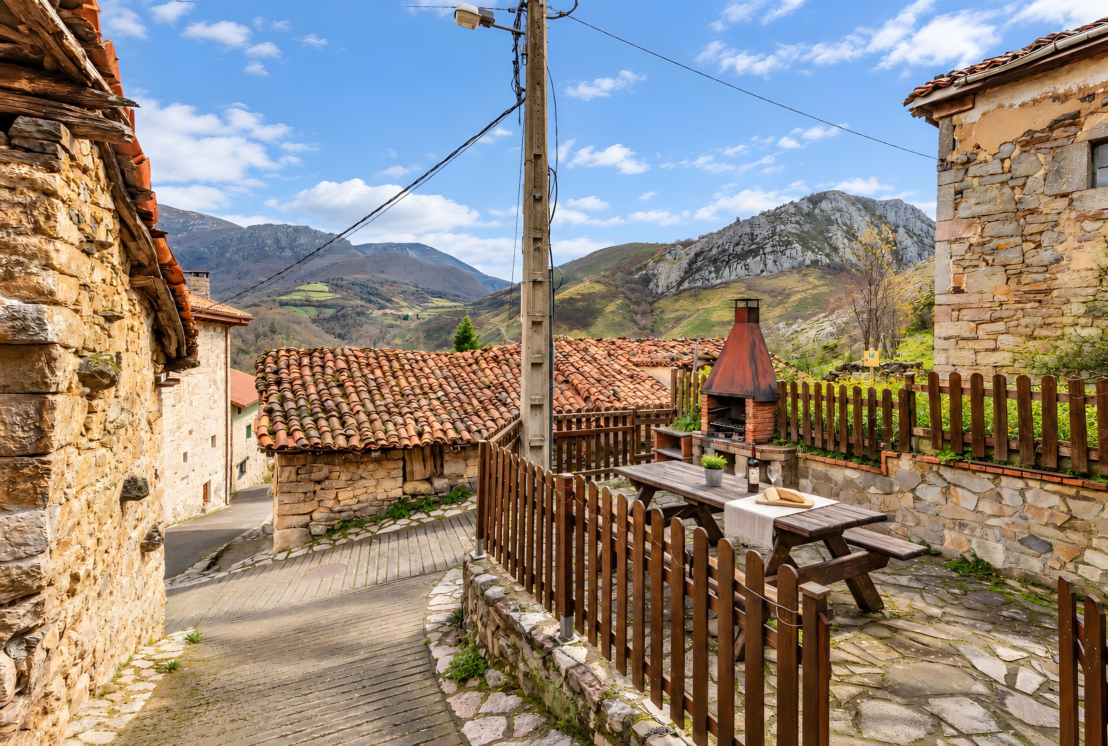 Un pueblo de piedra con tejados de teja, una valla de madera y un camino pavimentado que lleva a una mesa de madera con una botella de vino y una copa.
