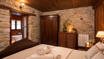 A bedroom with a stone wall, wooden ceiling, a bed with rolled towels, a wooden wardrobe, and a nightstand with a lamp.