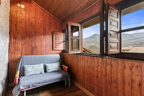 A wooden cabin interior with a sofa, a window with shutters, and a view of mountains.