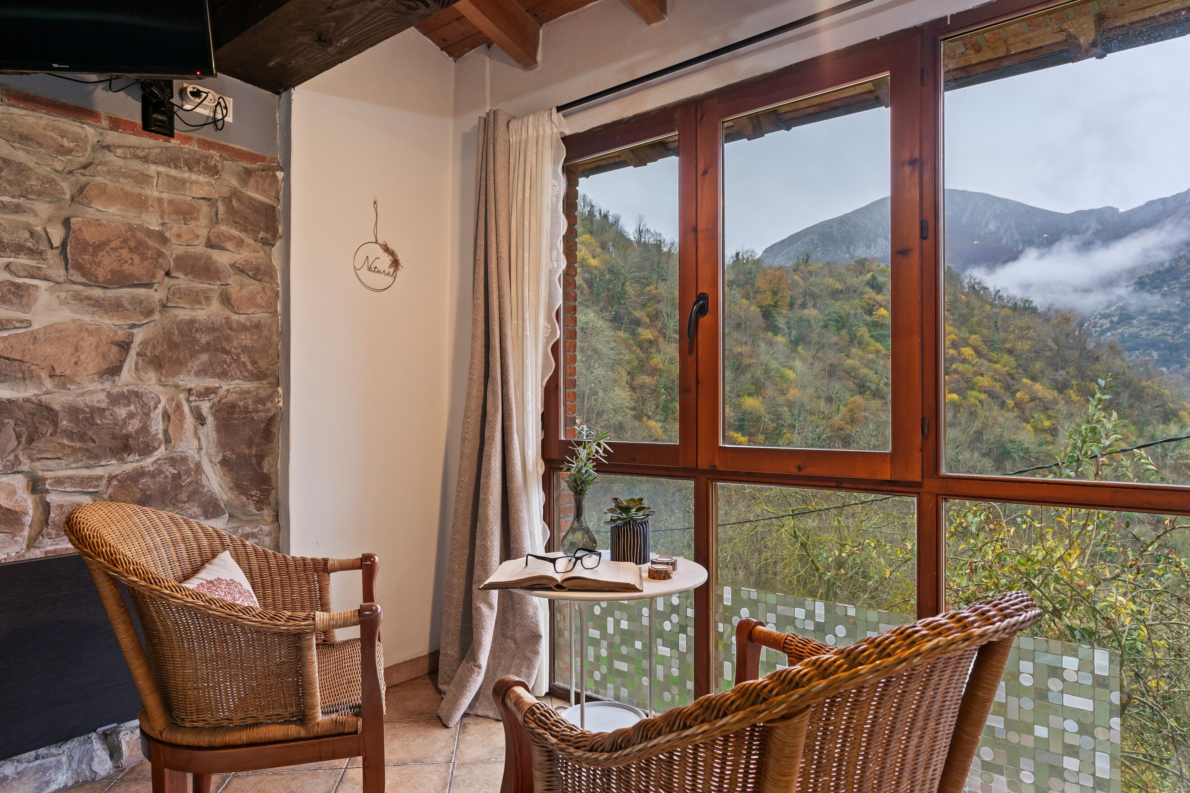 Una habitación con pared de piedra, techo de madera, dos sillas de mimbre, una mesita con un libro y vasos, y un ventanal con vistas a la montaña.