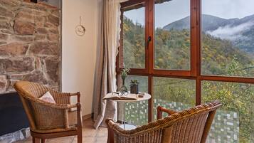 Una habitación con pared de piedra, techo de madera, dos sillas de mimbre, una mesita con un libro y vasos, y un ventanal con vistas a la montaña.