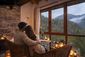 Ein Paar sitzt an einem Fenster mit Blick auf eine gebirgige Landschaft, mit einer Steinmauer und einem kleinen Tisch mit Kerzen und einem Buch.