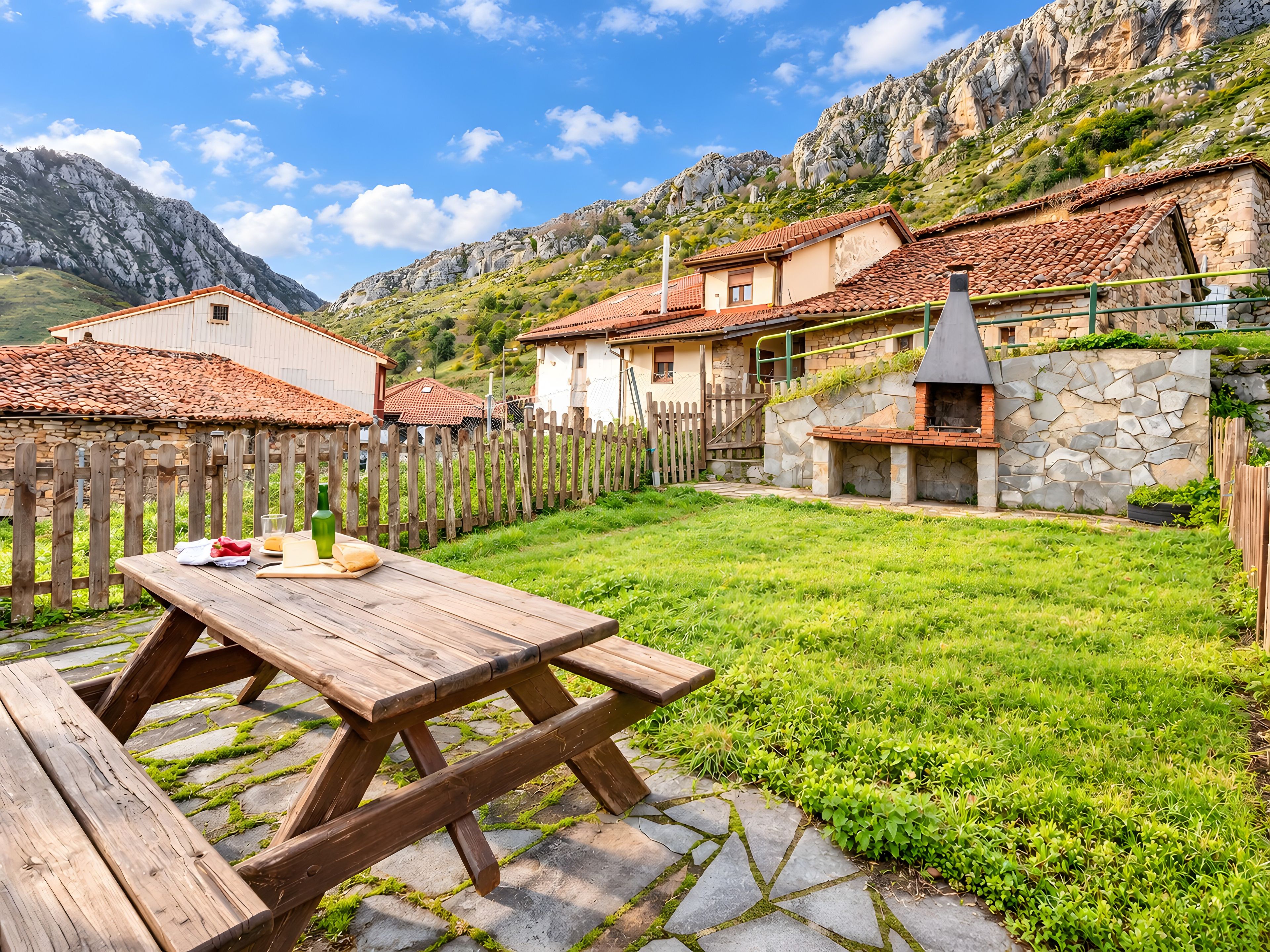 A wooden picnic table in a grassy backyard with stone flooring, surrounded by a wooden fence, and houses with red-tiled roofs in the background.